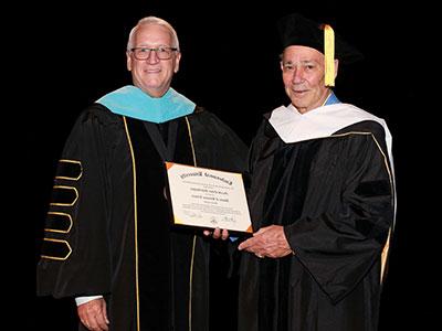 Jerry Scheidegger wearing black doctoral robes with white hood, being handed his honorary doctorate by President Porter wearing black doctoral robes with blue hood.